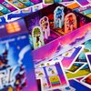 Na zdjęciu znajduje się gra planszowa "MUDUKO Hotel 60 zagadek" z kolorowymi elementami, w tym pionkami z postaciami i kartami z ilustracjami. Opakowanie gry jest widoczne z przodu, a w tle rozłożone są różnobarwne karty.