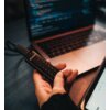 Na zdjęciu widoczny jest pendrive z klawiaturą numeryczną, trzymany w dłoni, z wyraźnym logo "Verbatim". W tle znajduje się otwarty laptop z widocznym ekranem, na którym wyświetlany jest kod programistyczny.
