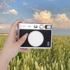 Na zdjęciu widać rękę trzymającą aparat z dekielkiem LOVEINSTANT SB7952 do Instax Mini Evo Srebrny, na tle pola zboża i nieba z chmurami. Na aparacie widoczne są napisy: "FUJIFILM", "INSTAX mini Evo" oraz "instax mini Evo".