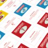 Na zdjęciu widoczne są karty z gry planszowej Monopoly Mega, z różnymi kolorami tła i napisami, takimi jak "MONOPOLY", "SZANSA", "KASA SPOŁECZNA" oraz ilustracjami, np. autobusu i domów. Teksty na kartach zawierają instrukcje gry, np. "NAJWIĘKSZA I NAJWIĘ