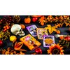 Na zdjęciu znajduje się gra karciana "Wirus! Halloween" w pomarańczowym pudełku z ilustracją postaci w stroju wampira. Wokół pudełka rozłożone są karty z motywami Halloween, czaszka, sztuczne dynie, liście i inne dekoracje w jesiennych kolorach.