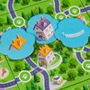 Plansza do gry przedstawia kolorową mapę z drogami, zielonymi terenami i budynkami, na której znajdują się niebieskie żetony z ilustracjami domów. Na mapie widoczne są także symbole łapek w białych okręgach.