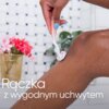 Na zdjęciu widoczna jest ręka trzymająca różową golarkę, która przesuwa się po nodze, a w tle znajduje się koszyk z różowymi kwiatami. Na zdjęciu widnieje tekst: "Rączka z wygodnym uchwytem".
