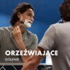 Mężczyzna w szarej koszulce goli się przed lustrem, używając maszynki do golenia i pianki. Na zdjęciu widoczny jest tekst: "ORZEŹWIAJĄCE GOLENIE".