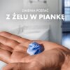 Na zdjęciu widoczna jest dłoń trzymająca niebiesko-białą piankę, a w tle znajduje się rozmyta łazienka. Na górze zdjęcia umieszczony jest tekst: "ZMIENIA POSTAĆ Z ŻELU W PIANKĘ".