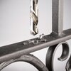 Wiertło metalowe jest umieszczone nad metalową powierzchnią, na której znajdują się opiłki metalu. W tle widoczna jest część metalowej konstrukcji z ozdobnym elementem.