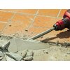 Na zdjęciu widoczny jest czerwony młot udarowy z dłutem, który rozbija jasnoszary beton na tle pomarańczowych płytek ceramicznych. Wokół dłuta znajdują się fragmenty pokruszonego betonu i pył.