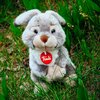 Pluszowy zając o szarym futerku siedzi na trawie, z czerwonym medalionem z napisem "trudi" na szyi. W tle widoczne są zielone liście i źdźbła trawy.