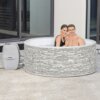 Dmuchane jacuzzi BESTWAY Lay-Z-Spa Vancouver AirJet Plus o wymiarach 155 x 60 cm, z szarym wzorem przypominającym kamień, stoi na tarasie przed dużymi oknami. Na boku jacuzzi widoczne jest logo "LAY-Z-SPA VANCOUVER".