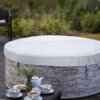 Dmuchane jacuzzi z szarym pokrowcem, na którym widnieje napis "LAY-Z-SPA VANCOUVER", stoi na drewnianym tarasie, a w tle widać zieleń ogrodu. Na pierwszym planie znajduje się stolik z białą zastawą, w tym filiżankami, dzbankiem i talerzykami z ciastkami.