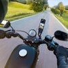 Zdjęcie przedstawia widok z perspektywy motocyklisty jadącego drogą, z zamontowaną na kierownicy kamerą marki INSTA360, która rejestruje trasę. W tle widać zielone pola i drzewa, a na ekranie kamery wyświetla się obraz drogi.