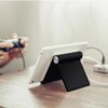 Czarna podstawka do tabletu UGREEN LP115 podtrzymuje biały tablet na drewnianym blacie. W tle widoczna jest osoba trzymająca kontroler do gier oraz kabel podłączony do tabletu.