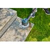 Na zdjęciu widoczna jest szczotka obrotowa czyszcząca powierzchnię kamiennych schodów, otoczona zieloną trawą. Na szczotce znajduje się logo "GARDENA".