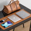 Na zdjęciu widać czarny stół z marmurowym blatem, na którym leży brązowa torba, szare etui na laptopa z napisem "UGREEN", srebrny laptop z logo jabłka, smartfon, tablet oraz zegarek na brązowym pasku. Wszystkie przedmioty są ułożone na brązowej podkładce.