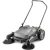 Na zdjęciu znajduje się szara zamiatarka ręczna z dwoma dużymi szczotkami z przodu i uchwytem do prowadzenia. Na obudowie widoczne jest logo "KARCHER".