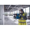 Na zdjęciu widać osobę w kasku, masce ochronnej i nausznikach, używającą młota udarowo-obrotowego BOSCH GBH 18V-28 CF do wiercenia w betonowej ścianie. W tle znajduje się przestrzeń budowlana z dużymi oknami i widoczną konstrukcją sufitu.