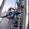 Osoba w kasku i ochronnikach słuchu używa młotowiertarki BOSCH PROFESSIONAL GBH 18V-34 CF do wiercenia w ścianie, w tle widoczne są rusztowania. Na urządzeniu widoczne jest logo BOSCH.