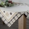 Beżowy obrus o wymiarach 90 x 90 cm z wzorem w śnieżynki i kratę, leży na drewnianym stole. Na obrusie znajduje się dekoracja z zielonych gałązek i czerwonych jagód.
