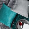Na zdjęciu widoczne są dwie poduszki w poszewkach, jedna w kolorze turkusowym, druga szara, leżące na wzorzystym materiale. Obok znajduje się okrągła taca z filiżanką herbaty i białą książką.