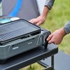 Na zdjęciu widoczny jest grill turystyczny z napisem "CAMPINGAZ Bistro Grill", ustawiony na stole, z ręką regulującą pokrętło. W tle znajduje się rozmyty krajobraz z trawą i fragmentem namiotu.