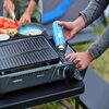 Osoba wkłada niebieską butlę gazową do turystycznego grilla CAMPINGAZ Camp Bistro Grill, który stoi na czarnym stole. W tle widać talerz z pokrojonymi pomidorami i innymi składnikami.