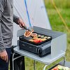 Osoba używa szczypiec do obracania jedzenia na grillu turystycznym CAMPINGAZ Camp Bistro Grill, na którym znajdują się kawałki mięsa oraz warzywa, takie jak papryka i cukinia. Grill stoi na stoliku na tle namiotu i trawy, a na jego froncie widoczne jest l