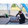 Wnętrze namiotu z widocznym materacem z napisem "Coleman Supercomfort 7.5 Double" oraz stolikiem turystycznym z kubkiem i lampą. W tle znajduje się plecak, a przez otwarte wejście namiotu widać trawę i drzewa.