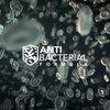 Na zdjęciu widać rozmyte, półprzezroczyste kształty przypominające komórki na ciemnym tle. Na pierwszym planie znajduje się biały napis "ANTI BACTERIAL FORMULA" obok symbolu tarczy z mikroorganizmami.