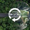 Zdjęcie przedstawia widok z lotu ptaka na drogę biegnącą przez gęsty las, z fragmentem Ziemi widocznym w tle. Na środku obrazu znajduje się biały symbol recyklingu z napisem "CONTAINS Recycled MATERIAL".