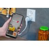 Na zdjęciu widać rękę trzymającą smartfon z aplikacją "Setti+ SMART HOME" na ekranie, z ikonami Google Play, App Store i Powered by Tuya. W tle znajduje się gniazdko elektryczne z podłączonym urządzeniem oraz słoik z marchewkami.