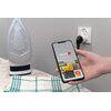 Na zdjęciu widać rękę trzymającą smartfon z aplikacją "Setti+ SMART HOME" na ekranie, z widocznymi logotypami Google Play i App Store oraz napisem "powered by tuya". W tle znajduje się żelazko stojące na desce do prasowania, a w ścianie widać gniazdko ele