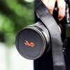 Na zdjęciu widoczny jest czarny dekielek magnetyczny z pomarańczowym logo na obiektywie aparatu, trzymanym przez osobę. W tle znajdują się rozmyte zielone liście.