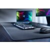 Na zdjęciu znajduje się klawiatura z podświetleniem RGB umieszczona na dużej podkładce, obok której leży mysz. W tle widoczne są dwa monitory z kolorowymi, abstrakcyjnymi wzorami na ekranach.