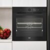 Czarny piekarnik elektryczny w zabudowie z wyświetlaczem pokazującym czas 00:35 i temperaturę 230°C, poniżej znajduje się logo "beko". Obok piekarnika stoi misa z czerwonymi jabłkami na blacie kuchennym.