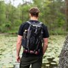 Na zdjęciu widoczny jest czarny plecak z napisem "NILS CAMP" oraz "Valley NC1749", noszony przez osobę stojącą nad stawem pokrytym liliami wodnymi. W bocznej kieszeni plecaka znajduje się czarna butelka z napisem "STNLS", a na dole plecaka przymocowany je