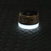 Na zdjęciu znajduje się brązowa lampa kempingowa z napisem "NILS" na ciemnym, teksturowanym tle. Lampa emituje jasne, białe światło u podstawy.