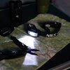 Na zdjęciu znajduje się latarka czołowa NILS CAMP NC0006, świecąca na rozłożoną mapę topograficzną. Obok latarki widoczne są narzędzia wielofunkcyjne oraz karabińczyk z napisem "NILS".