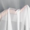 Na zdjęciu widoczna jest biała firana zawieszona na karniszu, która sięga do podłogi. W tle znajduje się jasne okno z widokiem na zieleń na zewnątrz.