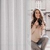 Na zdjęciu widoczna jest biała zasłona w drobne kropki, obok której siedzi osoba trzymająca czarny kubek. W tle znajduje się rozmyty widok na budynek z dużymi oknami.