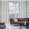 Na zdjęciu znajduje się salon z dużymi oknami, przez które widać balkon z ozdobną balustradą i widokiem na miasto. W pomieszczeniu znajduje się fioletowa sofa z kolorowymi poduszkami, szklany stolik z dekoracjami oraz białe zasłony w delikatny wzór.