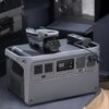 Na zdjęciu znajduje się szary generator przenośny z wyświetlaczem pokazującym "100" oraz różnymi portami i przyciskami. Na górze generatora umieszczony jest złożony dron, a obok niego leży kabel z podłączonym urządzeniem.