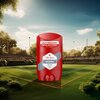 Czerwony dezodorant w sztyfcie z napisem "24/7 FRESSSHHH PERFUME QUALITY SCENT" oraz "Old Spice ORIGINAL RICH SCENT DEODORANT STICK" stoi na tle kortu tenisowego otoczonego drzewami. W tle widoczne są trybuny i maszty oświetleniowe.