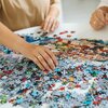 Na zdjęciu widać ręce układające puzzle na białym stole, z widocznymi fragmentami kolorowego obrazka przedstawiającego miasto. Puzzle są w trakcie układania, a wokół leży wiele nieułożonych elementów.