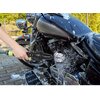 Na zdjęciu widoczna jest ręka trzymająca szczotkę do felg z logo "DUNLOP", która myje motocykl pokryty pianą. W tle znajduje się zieleń i brukowana nawierzchnia.