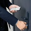 Osoba trzyma smartfon z aplikacją pokazującą zieloną ikonę kłódki i napis "UNLOCKED", jednocześnie przekręcając gałkę zamka w drzwiach. Drzwi są ciemnoszare, a klamka i zamek mają nowoczesny design.