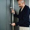 Mężczyzna w ciemnym swetrze trzyma duży, metalowy uchwyt drzwi, wkładając klucz do zamka. Drzwi są w kolorze szarym, z poziomymi panelami.