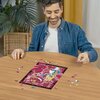 Na zdjęciu widać osobę układającą puzzle na okrągłym stole, z widocznym obrazkiem przedstawiającym postacie z "Alicji w Krainie Czarów" na tle różowych i czerwonych odcieni. W tle znajduje się roślina doniczkowa oraz zasłona.