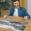 Na zdjęciu widać osobę układającą puzzle przedstawiające samochód Porsche 911R, z kilkoma brakującymi elementami na drewnianym stole. W tle znajduje się roślina doniczkowa oraz zasłony.