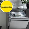 Otwarta zmywarka z widocznymi naczyniami w środku, na górze znajduje się stos talerzy i kieliszki, a obok roślina doniczkowa. Na żółtym tle widnieje napis: "AUTOMATYCZNE OTWIERANIE DRZWI".