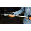 Osoba w rękawiczkach trzyma metalową pompę do oleju z widocznym napisem "NEO TOOLS" na cylindrze. Pompa ma pomarańczowy uchwyt i jest wykonana z błyszczącego metalu.